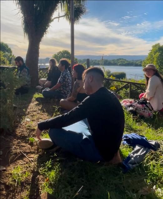 Público sentado en el pasto del patio mirando el río al atardecer